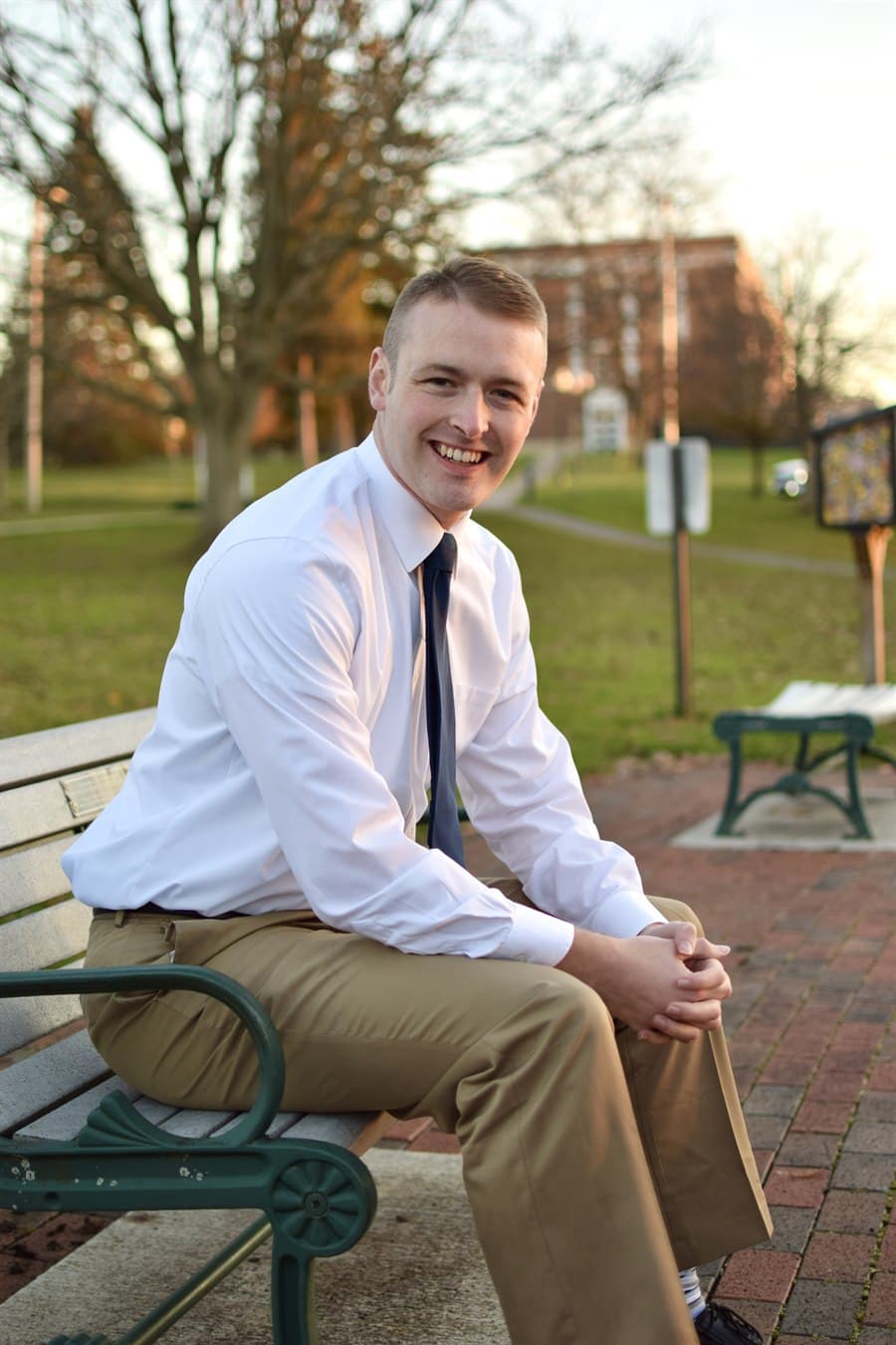 James Corl, seated on a park bench in a white shirt and tie.