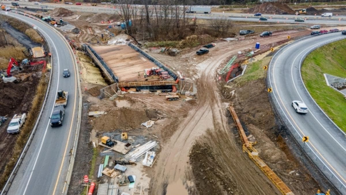 Aerial view of highway interchange construction: a new bridge deck under build between two curving carriageways, with excavators, materials and churned earth around it.